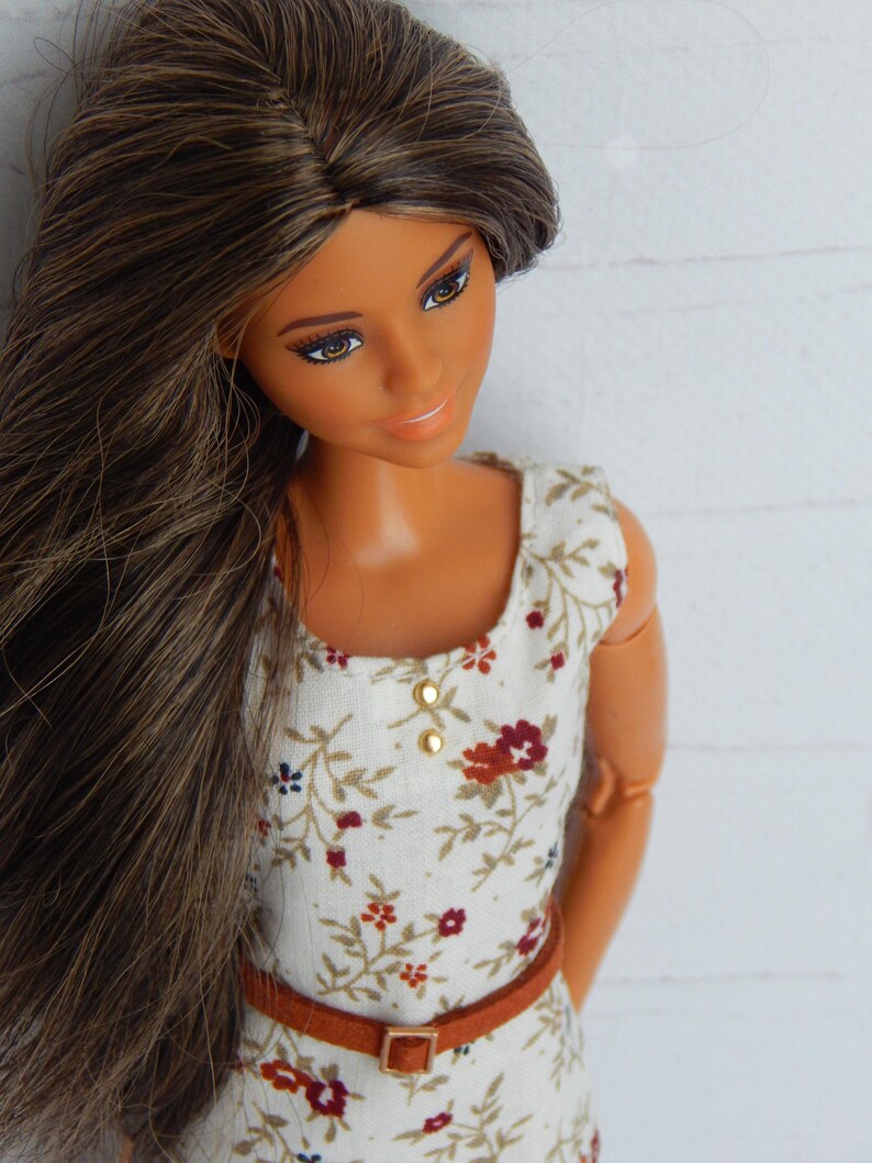 K&ouml;nnte beinhalten: Eine Puppe mit langen braunen Haaren, die ein wei&szlig;es Kleid mit Blumenmuster und einen braunen G&uuml;rtel tr&auml;gt.