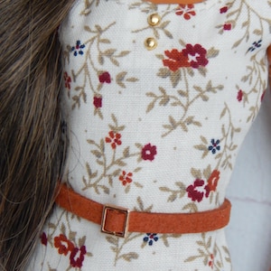 K&ouml;nnte beinhalten: Ein wei&szlig;es Kleid mit Blumenmuster und einem braunen G&uuml;rtel. Das Kleid hat einen runden Ausschnitt und kurze &Auml;rmel. Der G&uuml;rtel hat eine goldene Schnalle.