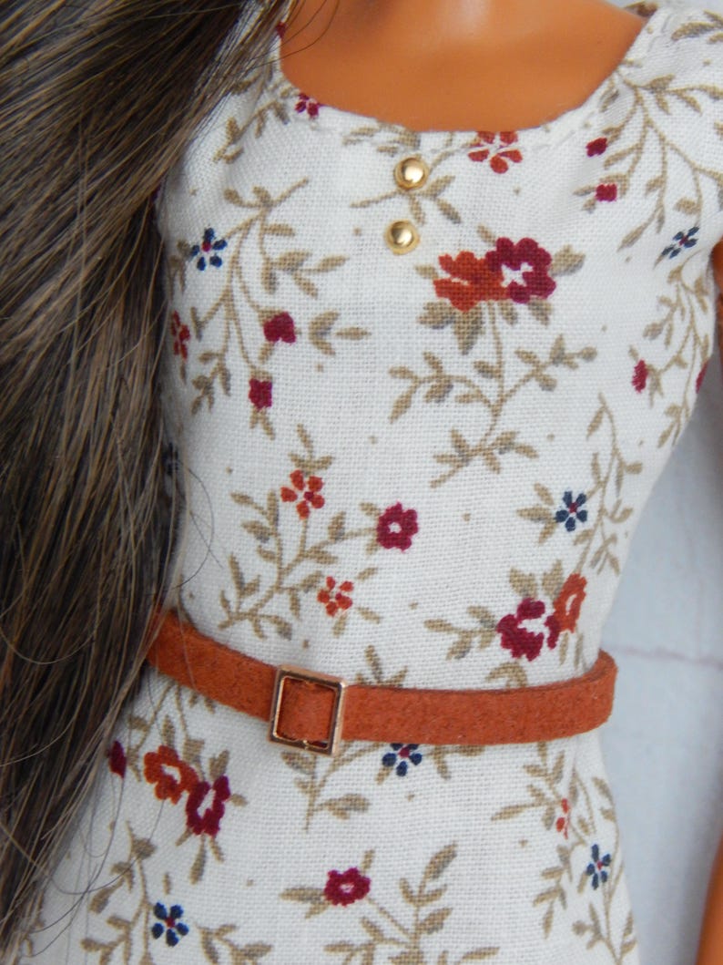 K&ouml;nnte beinhalten: Ein wei&szlig;es Kleid mit Blumenmuster und einem braunen G&uuml;rtel. Das Kleid hat einen runden Ausschnitt und kurze &Auml;rmel. Der G&uuml;rtel hat eine goldene Schnalle.