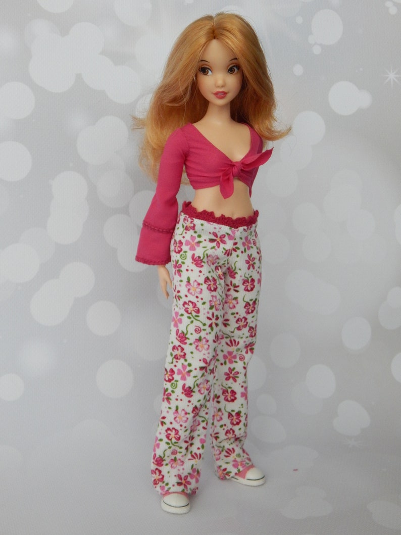May include: A doll with long, wavy, light brown hair wearing a pink crop top with a tie at the front and white pants with a pink floral pattern.