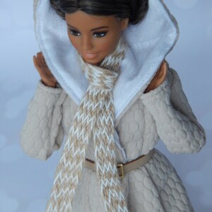 Puede incluir: Una mu&ntilde;eca con un abrigo de punto beige con capucha blanca y una bufanda marr&oacute;n y blanca. La mu&ntilde;eca tiene el pelo casta&ntilde;o y los ojos marrones.