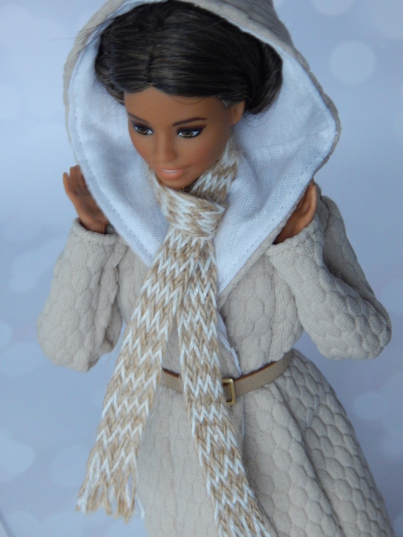 Puede incluir: Una mu&ntilde;eca con un abrigo de punto beige con capucha blanca y una bufanda marr&oacute;n y blanca. La mu&ntilde;eca tiene el pelo casta&ntilde;o y los ojos marrones.
