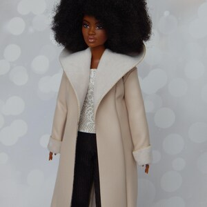 May include: A doll with an afro hairstyle wearing a beige coat with a white fur collar and cuffs. The doll is wearing a white lace top and black pants.
