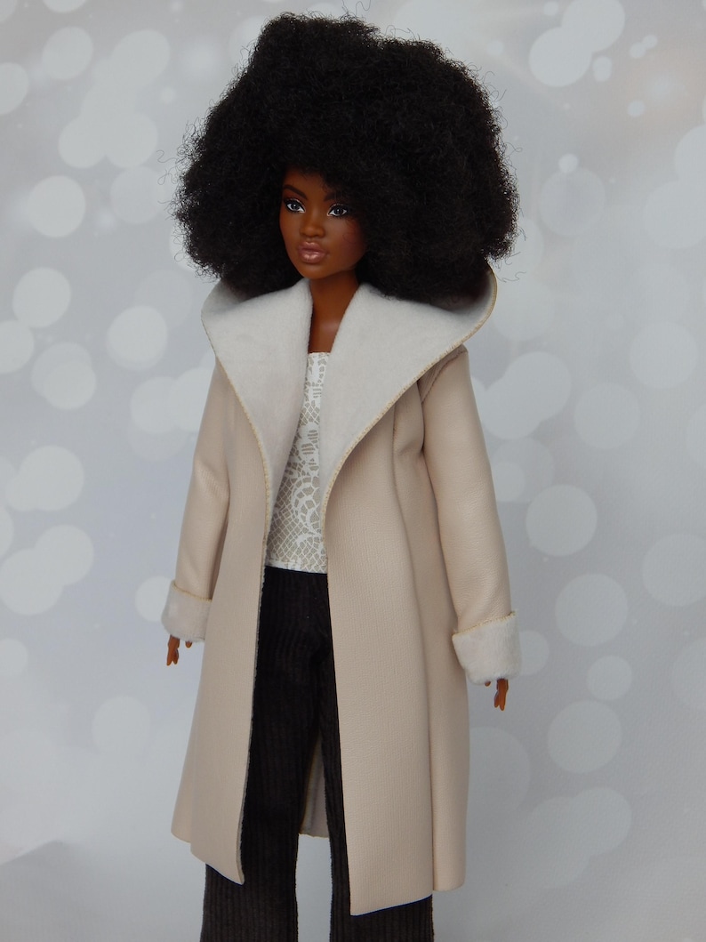 May include: A doll with an afro hairstyle wearing a beige coat with a white fur collar and cuffs. The doll is wearing a white lace top and black pants.
