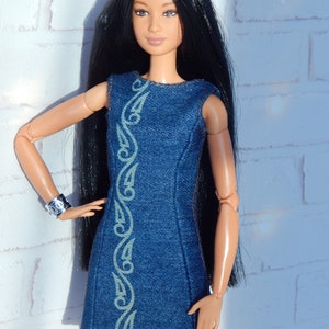May include: A doll wearing a blue denim dress with a decorative pattern. The dress has a sleeveless design and a straight cut. The doll has long black hair and brown eyes.