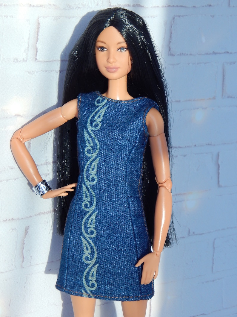 May include: A doll wearing a blue denim dress with a decorative pattern. The dress has a sleeveless design and a straight cut. The doll has long black hair and brown eyes.