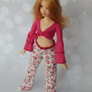 May include: A doll wearing a pink crop top with a tie at the front and white pants with a floral pattern. The doll has long, wavy, red hair.