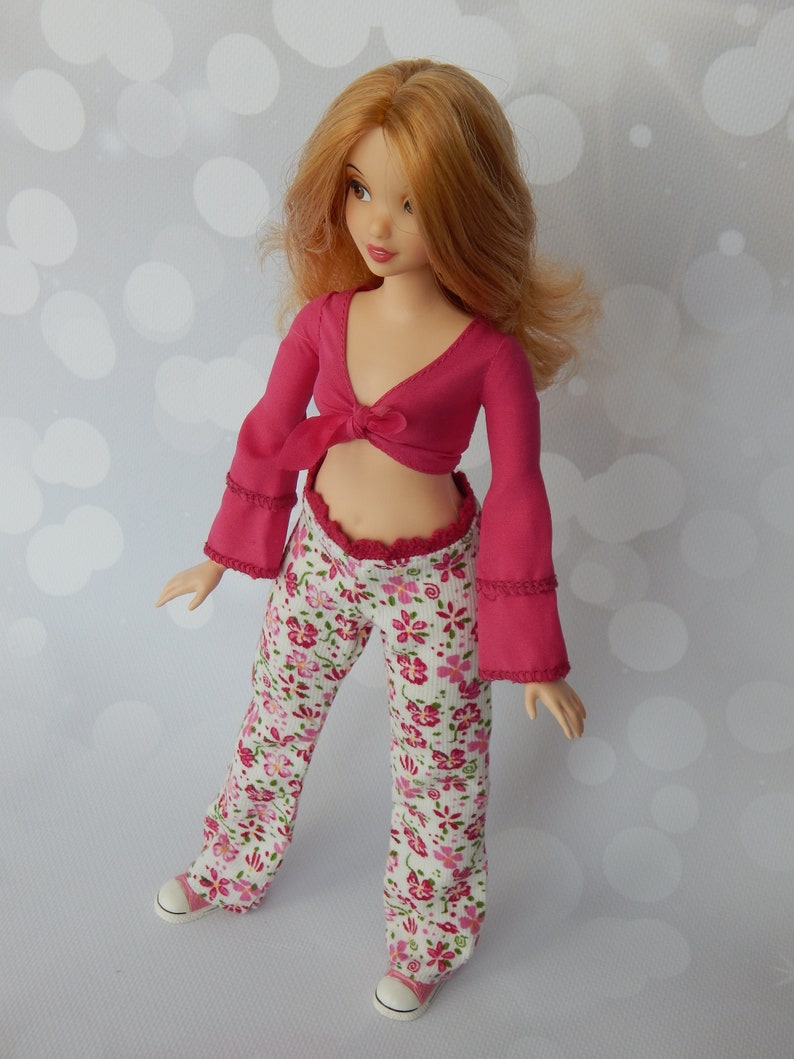 May include: A doll wearing a pink crop top with a tie at the front and white pants with a floral pattern. The doll has long, wavy, red hair.