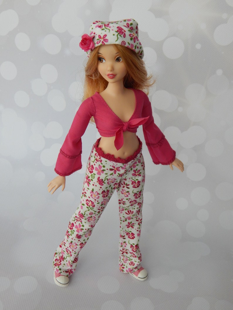 May include: A doll wearing a pink and white floral print outfit. The doll has long red hair and is wearing a white hat with pink flowers. The doll is wearing a pink crop top with a white floral print pants. The doll is wearing white sneakers.