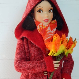 K&ouml;nnte beinhalten: Ein roter Kapuzenmantel mit einer flauschigen Textur und goldenen Kn&ouml;pfen. Der Mantel wird von einer Puppe mit schwarzen Haaren und einem roten Schal mit Blumenmuster getragen. Die Puppe h&auml;lt einen Strau&szlig; orangefarbener und gelber Blumen.