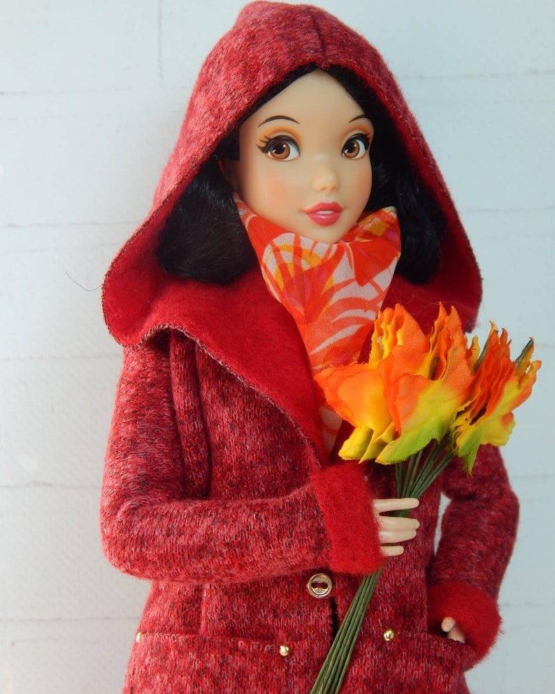 K&ouml;nnte beinhalten: Ein roter Kapuzenmantel mit einer flauschigen Textur und goldenen Kn&ouml;pfen. Der Mantel wird von einer Puppe mit schwarzen Haaren und einem roten Schal mit Blumenmuster getragen. Die Puppe h&auml;lt einen Strau&szlig; orangefarbener und gelber Blumen.