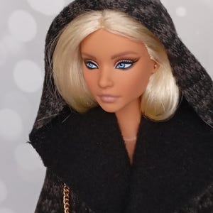 May include: A fashion doll with blonde hair and blue eyes, wearing a black hooded coat with a textured pattern. The doll has a fair complexion and is looking slightly to the side. A gold chain strap is visible.