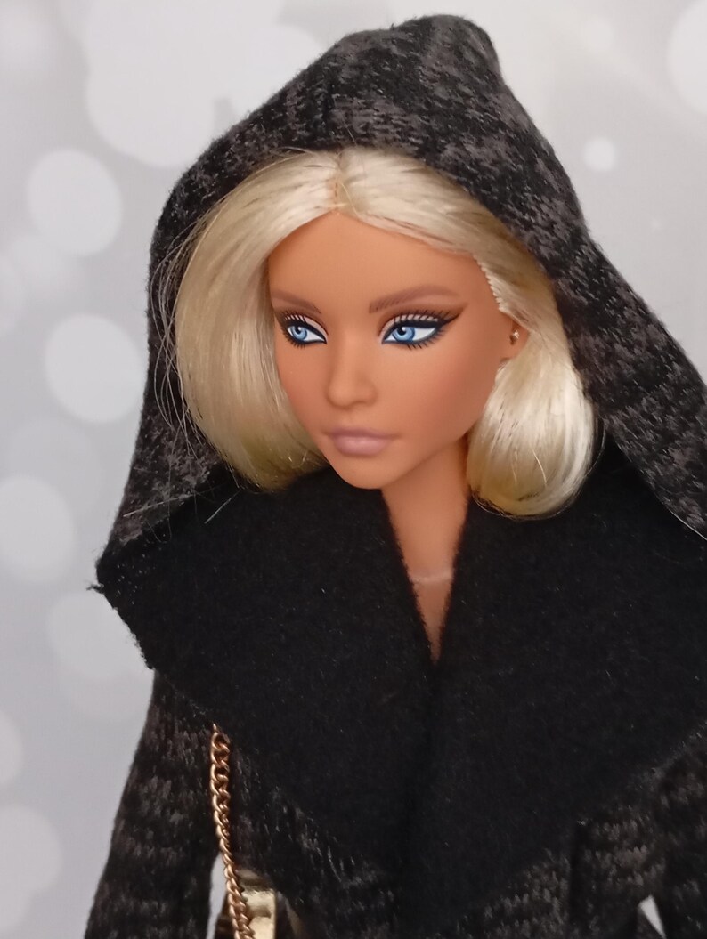 May include: A fashion doll with blonde hair and blue eyes, wearing a black hooded coat with a textured pattern. The doll has a fair complexion and is looking slightly to the side. A gold chain strap is visible.