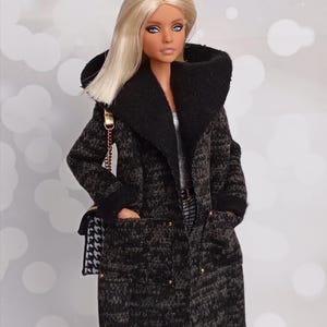 May include: A doll with blonde hair and blue eyes wearing a black and gray patterned coat with a large collar. The coat has gold buttons and pockets. A black and white purse with a chain strap hangs from the doll's shoulder.