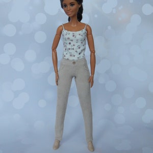 May include: A doll with long dark hair wearing a white floral tank top and beige corduroy pants.