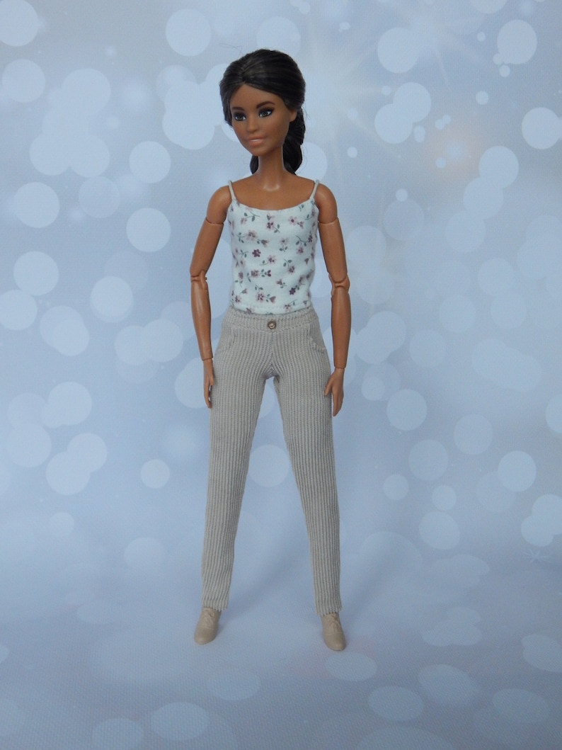 May include: A doll with long dark hair wearing a white floral tank top and beige corduroy pants.