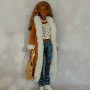 May include: A doll wearing a brown corduroy coat with a white fur trim, a white floral top, blue jeans with a distressed look, and white sneakers. The doll is holding a gold handbag and wearing yellow sunglasses.