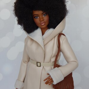 May include: A doll with dark brown skin and a large afro hairstyle is wearing a cream-colored faux fur coat with a brown leather purse.