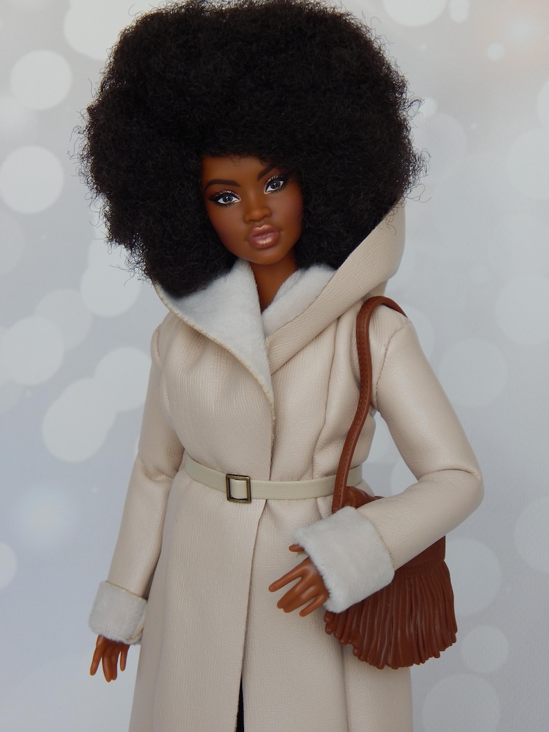 May include: A doll with dark brown skin and a large afro hairstyle is wearing a cream-colored faux fur coat with a brown leather purse.