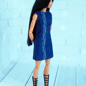 May include: A doll wearing a blue denim dress with a white embroidered design. The doll has long black hair and is wearing black gladiator sandals.