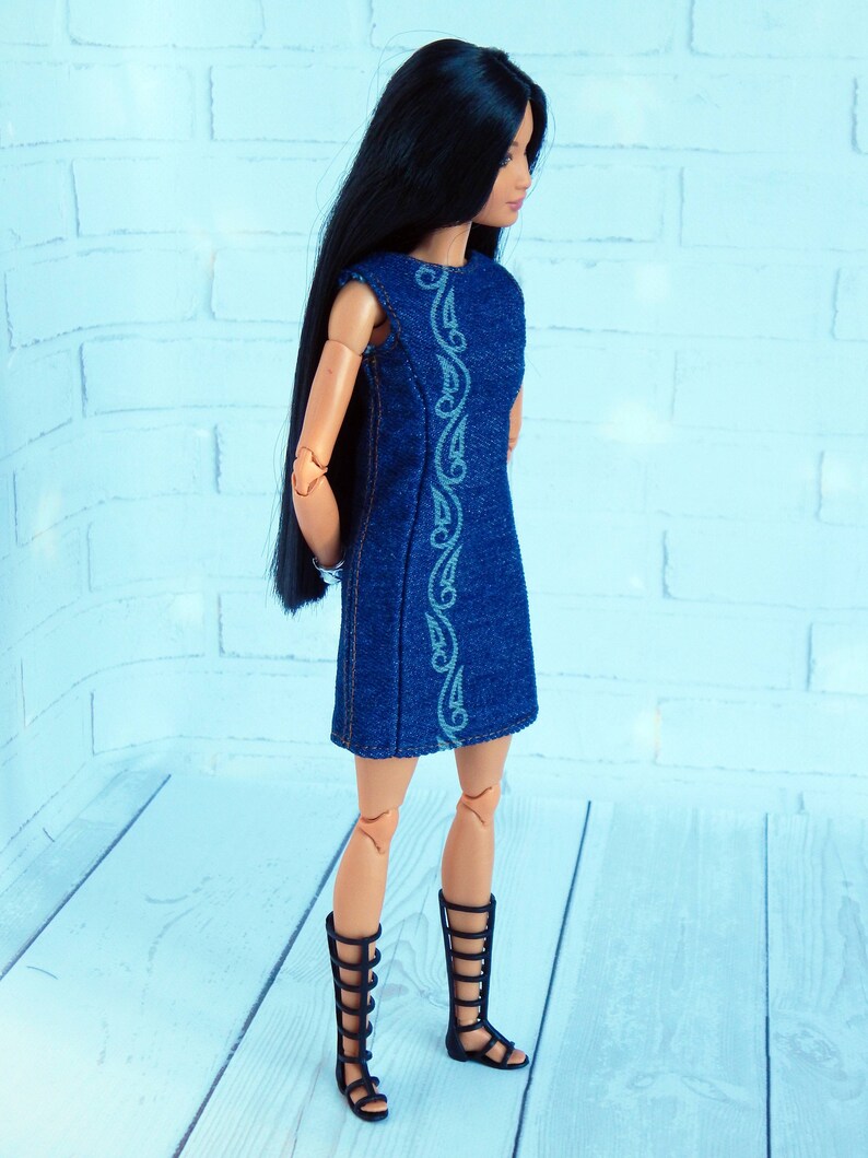 May include: A doll wearing a blue denim dress with a white embroidered design. The doll has long black hair and is wearing black gladiator sandals.