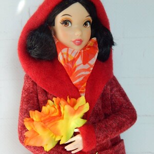 K&ouml;nnte beinhalten: Ein roter Kapuzenmantel mit flauschiger Textur und einem Blumenschal. Die Puppe h&auml;lt einen Strau&szlig; orangefarbener und gelber Blumen.