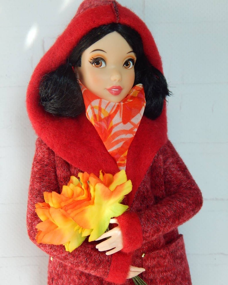 K&ouml;nnte beinhalten: Ein roter Kapuzenmantel mit flauschiger Textur und einem Blumenschal. Die Puppe h&auml;lt einen Strau&szlig; orangefarbener und gelber Blumen.