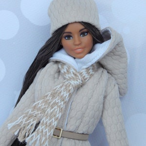 Puede incluir: Una mu&ntilde;eca con un abrigo de punto beige con ribete de piel blanca, una bufanda de punto beige y blanca y un gorro de punto beige con un pomp&oacute;n blanco.
