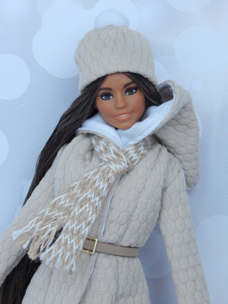 Puede incluir: Una mu&ntilde;eca con un abrigo de punto beige con ribete de piel blanca, una bufanda de punto beige y blanca y un gorro de punto beige con un pomp&oacute;n blanco.
