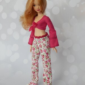 May include: A doll with long, wavy, red hair wearing a pink crop top with a bow and white pants with a floral pattern.