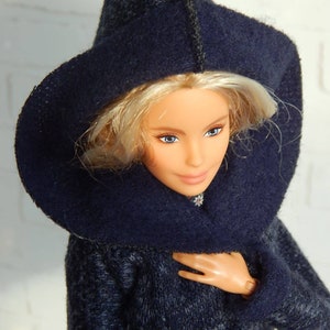 Pode incluir: Uma boneca vestindo um casaco azul escuro com capuz e textura felpuda. A boneca tem cabelo loiro e olhos azuis.