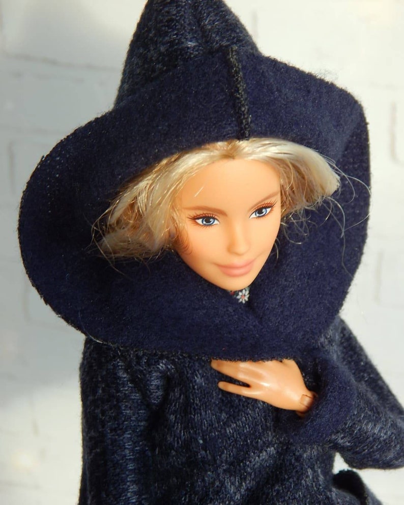 Pode incluir: Uma boneca vestindo um casaco azul escuro com capuz e textura felpuda. A boneca tem cabelo loiro e olhos azuis.