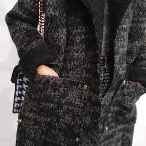 May include: A dark gray and black textured coat with a black collar and gold buttons. The coat has pockets and is paired with a small purse with a gold chain strap. The outfit includes a silver top and a black and white patterned skirt.