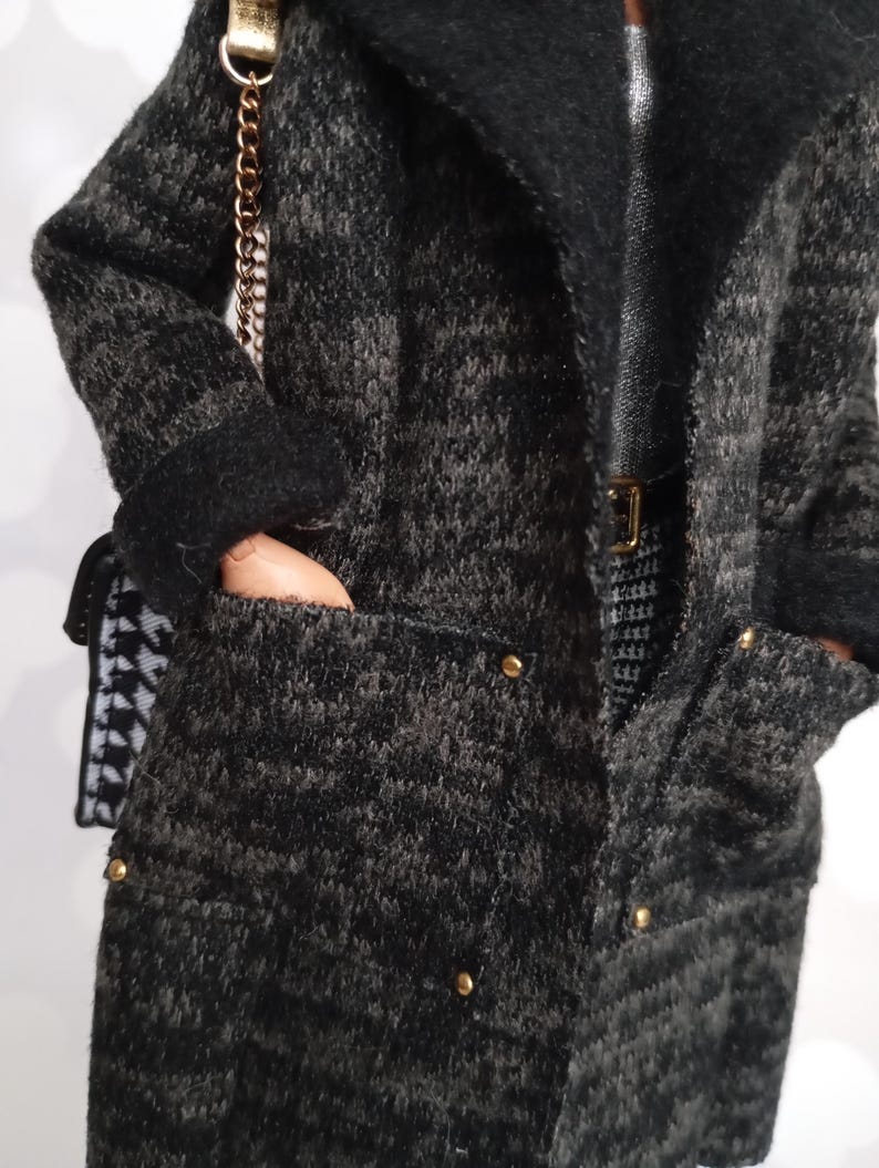 May include: A dark gray and black textured coat with a black collar and gold buttons. The coat has pockets and is paired with a small purse with a gold chain strap. The outfit includes a silver top and a black and white patterned skirt.