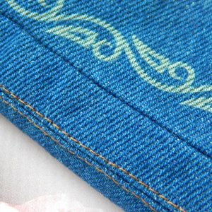 May include: Close-up of a blue denim fabric with a white patterned design. The fabric has a double stitched seam along the edge.