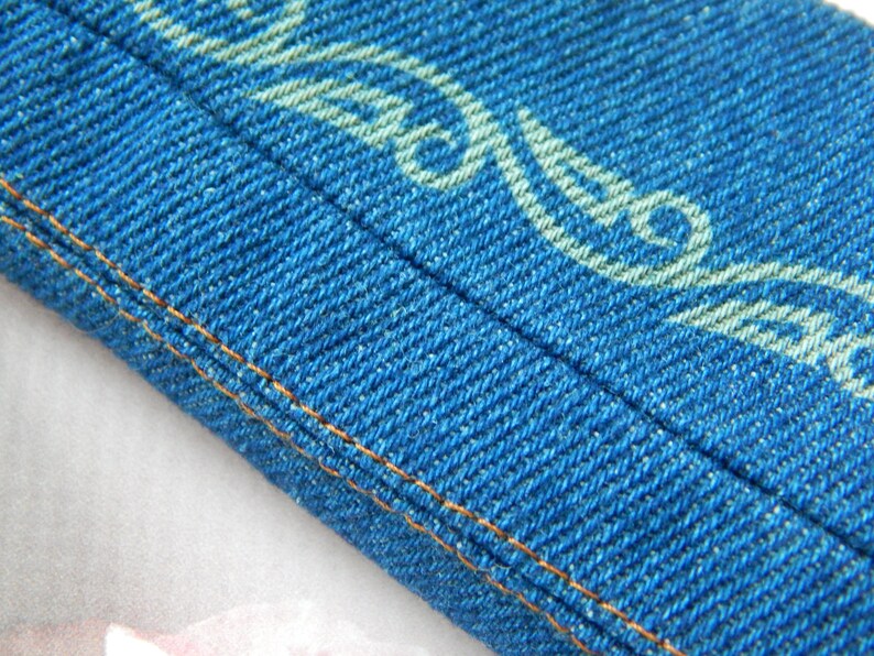May include: Close-up of a blue denim fabric with a white patterned design. The fabric has a double stitched seam along the edge.
