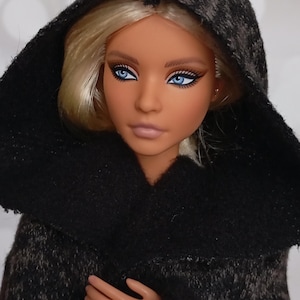 May include: A fashion doll with blonde hair and striking blue eyes, wearing a dark gray hooded cloak. The doll has a tan complexion and is wearing a gold ring. The cloak has a textured appearance.