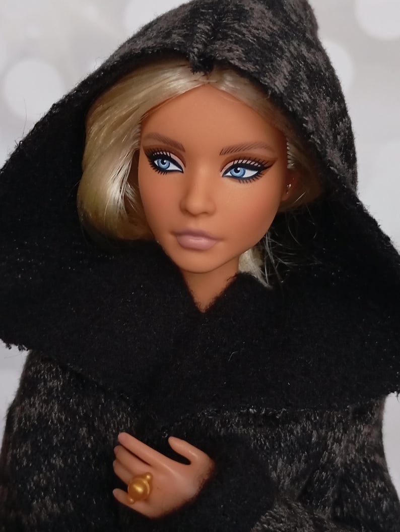 May include: A fashion doll with blonde hair and striking blue eyes, wearing a dark gray hooded cloak. The doll has a tan complexion and is wearing a gold ring. The cloak has a textured appearance.