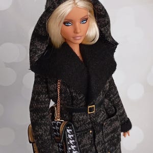 May include: A doll wearing a black and gray hooded coat with a black belt and a gold buckle. The coat has a black collar and a matching purse with a gold chain. The doll has blonde hair and blue eyes.