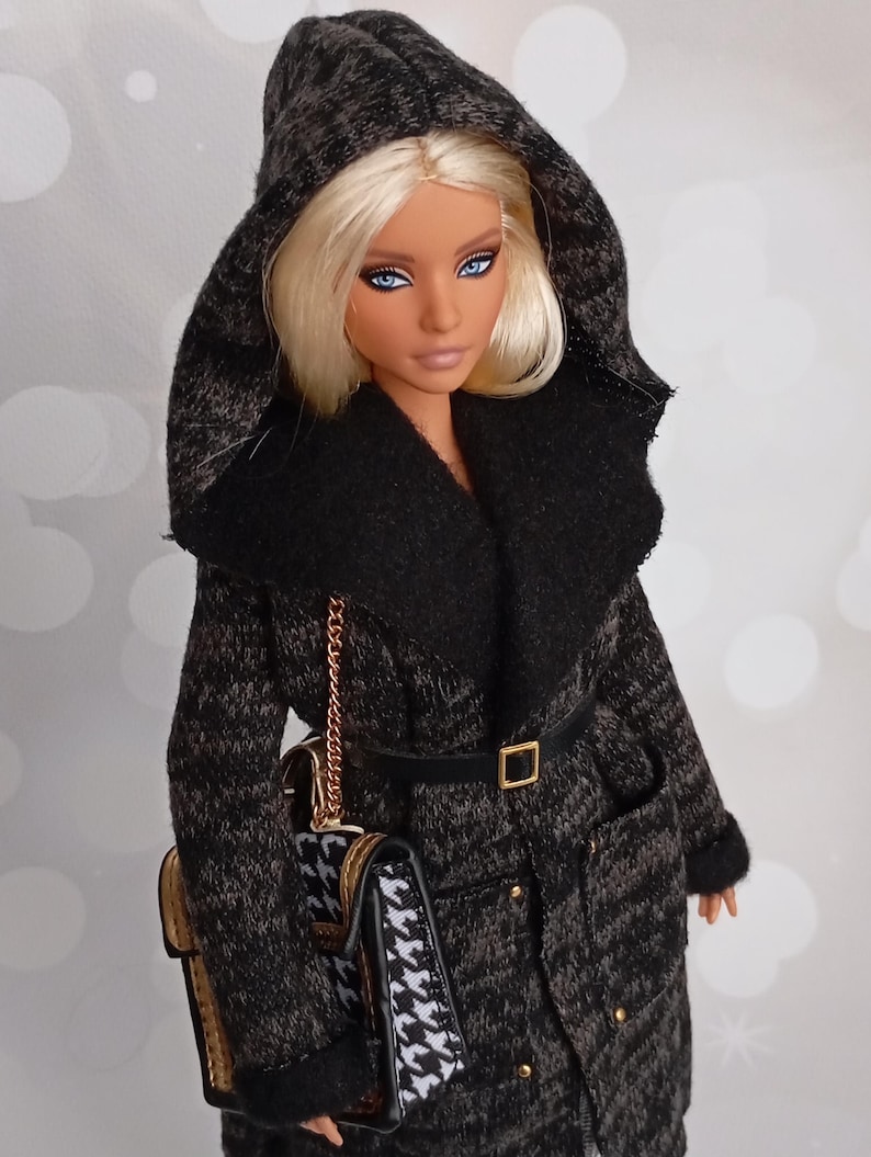May include: A doll wearing a black and gray hooded coat with a black belt and a gold buckle. The coat has a black collar and a matching purse with a gold chain. The doll has blonde hair and blue eyes.