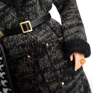 May include: A black and gray patterned coat with a black belt and gold buckle. The coat has gold buttons and black fur trim on the collar and cuffs. A gold ring is on the doll's finger.