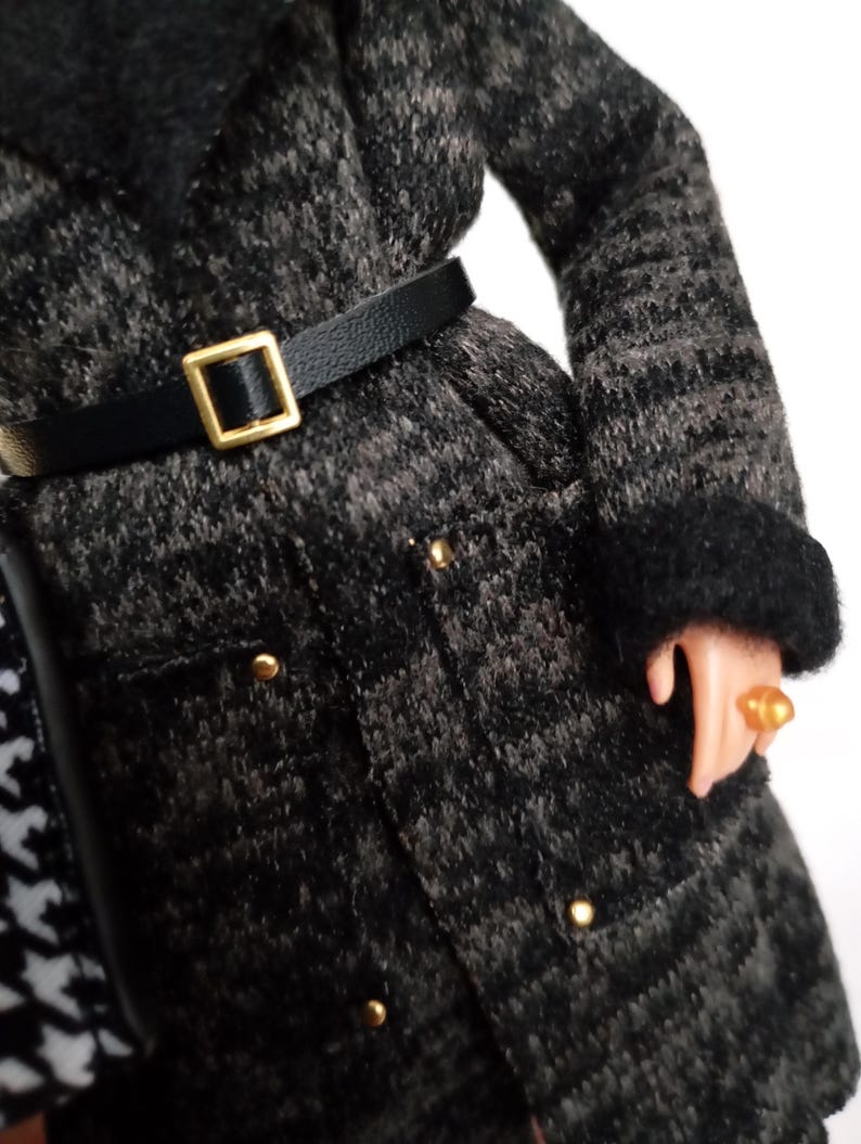 May include: A black and gray patterned coat with a black belt and gold buckle. The coat has gold buttons and black fur trim on the collar and cuffs. A gold ring is on the doll's finger.