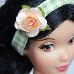 Puede incluir: Una muñeca con cabello negro que lleva una diadema verde y blanca con una flor de color melocotón. La muñeca tiene ojos marrones y lleva un vestido blanco.