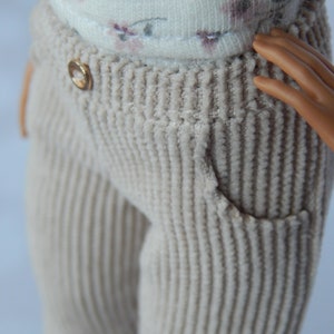 May include: Close-up of a pair of beige corduroy pants with a gold button. The pants have a ribbed texture and a single front pocket.