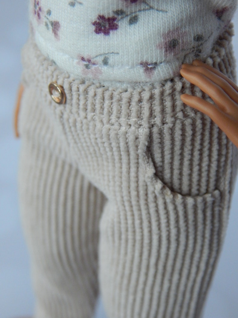 May include: Close-up of a pair of beige corduroy pants with a gold button. The pants have a ribbed texture and a single front pocket.