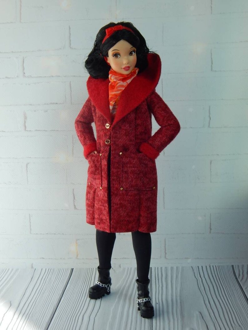 K&ouml;nnte beinhalten: Ein roter Wollmantel mit einem gro&szlig;en Kragen und goldenen Kn&ouml;pfen. Der Mantel wird von einer Puppe mit schwarzen Haaren und einem roten Stirnband getragen. Die Puppe tr&auml;gt schwarze Strumpfhosen und schwarze Stiefel mit silbernen Ketten.