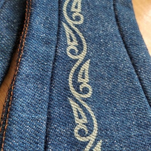 May include: A close-up of a blue denim fabric with a white decorative pattern. The fabric has a textured surface and is sewn with orange thread.