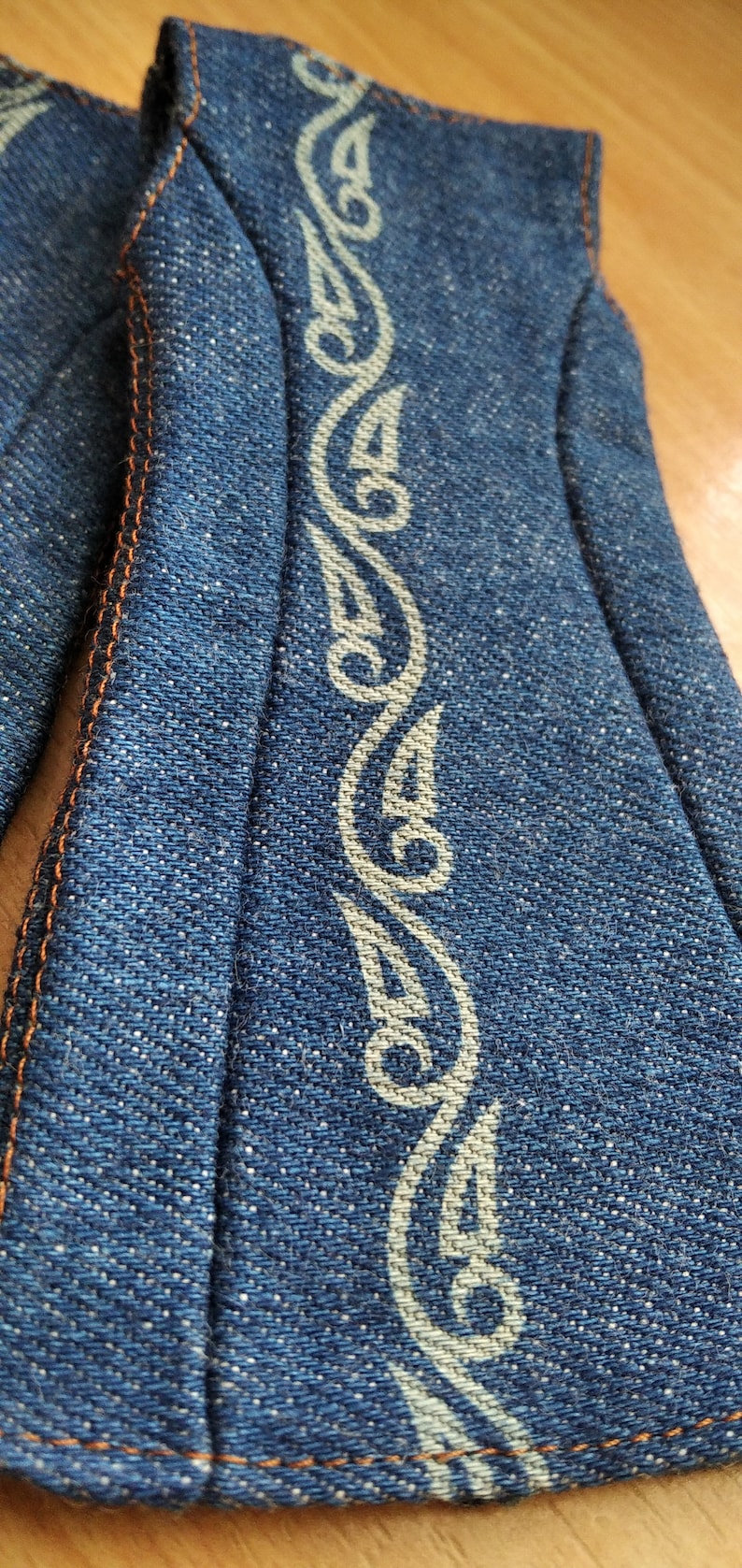 May include: A close-up of a blue denim fabric with a white decorative pattern. The fabric has a textured surface and is sewn with orange thread.