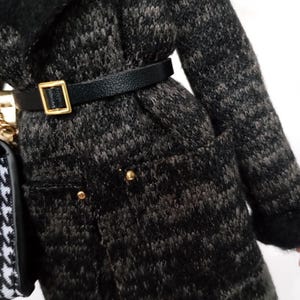 May include: A black and gray patterned coat with a black belt and gold buckle. The coat has gold buttons and a black collar. A black and white patterned purse is visible on the left side.