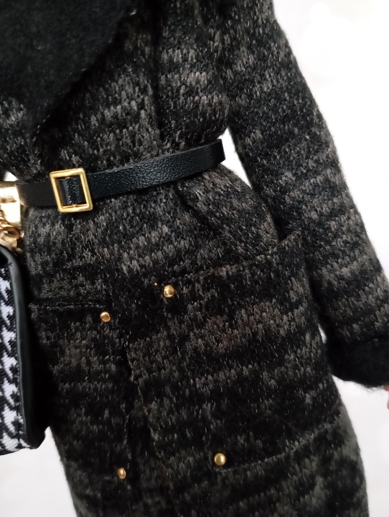 May include: A black and gray patterned coat with a black belt and gold buckle. The coat has gold buttons and a black collar. A black and white patterned purse is visible on the left side.